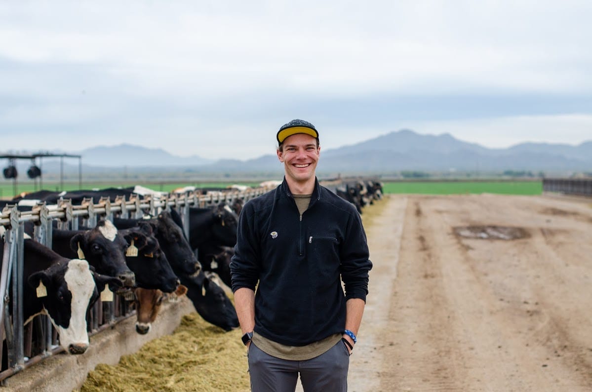 A person is standing and smiling at the camera while there are black cows in the background